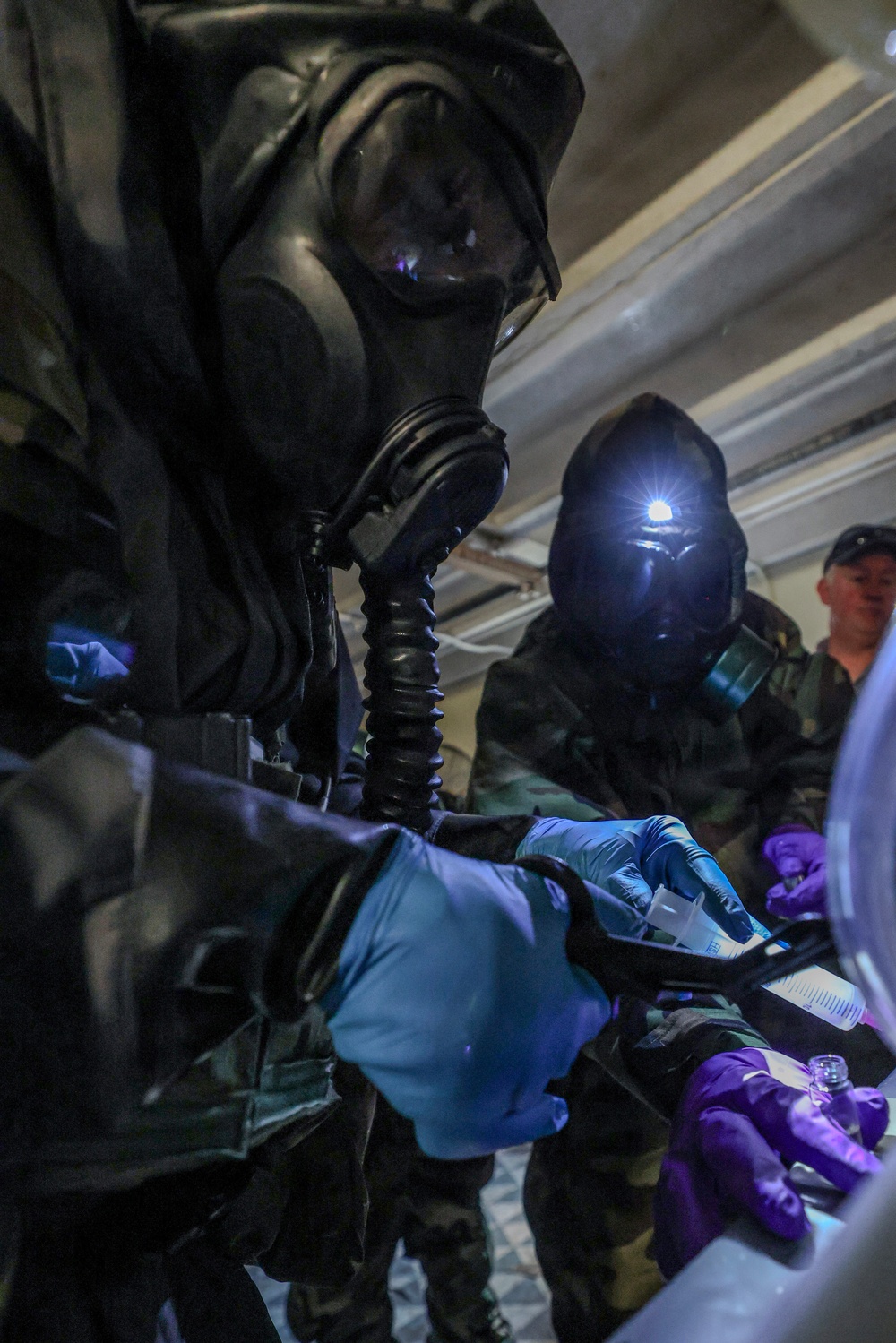 Two individuals wearing full CBRN (Chemical, Biological, Radiological, Nuclear) protective gear including gas masks and protective suits are conducting what appears to be a hazardous material sampling or testing procedure. They are wearing colored nitrile gloves (blue and purple) and working with small vials or test kits on a dark case or tray. A third person in the background, not wearing a gas mask, appears to be observing.