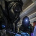 Two individuals wearing full CBRN (Chemical, Biological, Radiological, Nuclear) protective gear including gas masks and protective suits are conducting what appears to be a hazardous material sampling or testing procedure. They are wearing colored nitrile gloves (blue and purple) and working with small vials or test kits on a dark case or tray. A third person in the background, not wearing a gas mask, appears to be observing.