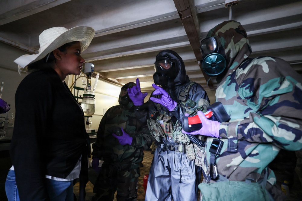 The image shows a woman in civilian clothing (white hat, black jacket) interacting with multiple personnel wearing military camouflage uniforms, gas masks, and purple nitrile gloves in what appears to be an indoor industrial or laboratory setting. The uniformed personnel appear to be conducting a CBRN (Chemical, Biological, Radiological, Nuclear) inspection or training exercise. Equipment including detection devices and protective gear is visible.