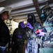 The image shows a woman in civilian clothing (white hat, black jacket) interacting with multiple personnel wearing military camouflage uniforms, gas masks, and purple nitrile gloves in what appears to be an indoor industrial or laboratory setting. The uniformed personnel appear to be conducting a CBRN (Chemical, Biological, Radiological, Nuclear) inspection or training exercise. Equipment including detection devices and protective gear is visible.