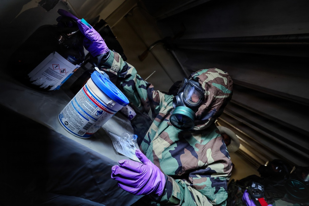 A person wearing military camouflage uniform, a full-face gas mask, and purple nitrile gloves is handling a cylindrical container with a blue lid in what appears to be a confined or darkened space. The individual appears to be conducting a decontamination, hazmat, or CBRN (Chemical, Biological, Radiological, Nuclear) response procedure. The setting and protective equipment suggest a military training exercise or operational response scenario.