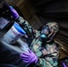 A person wearing military camouflage uniform, a full-face gas mask, and purple nitrile gloves is handling a cylindrical container with a blue lid in what appears to be a confined or darkened space. The individual appears to be conducting a decontamination, hazmat, or CBRN (Chemical, Biological, Radiological, Nuclear) response procedure. The setting and protective equipment suggest a military training exercise or operational response scenario.