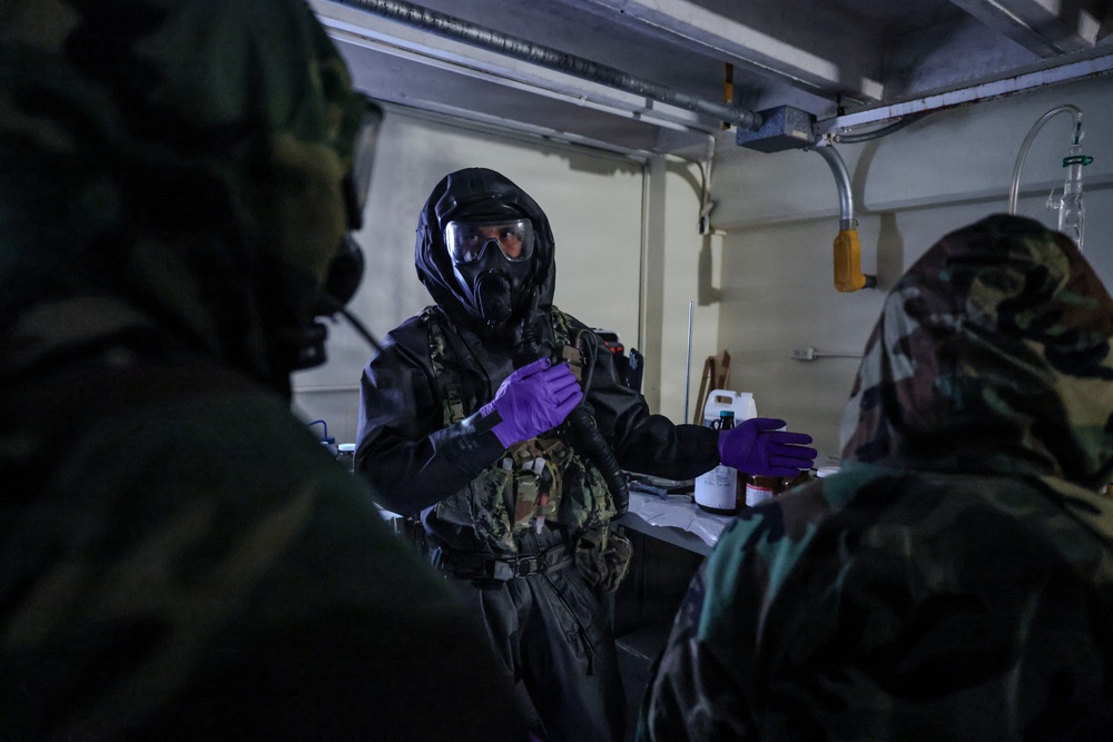 Military personnel are conducting what appears to be a CBRN (Chemical, Biological, Radiological, Nuclear) training exercise or operation in an enclosed workspace. The central figure is wearing full protective gear including a gas mask, black protective suit, and purple nitrile gloves, while interacting with others in camouflage uniforms. Chemical containers and laboratory equipment are visible on a workbench in the background.