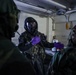 Military personnel are conducting what appears to be a CBRN (Chemical, Biological, Radiological, Nuclear) training exercise or operation in an enclosed workspace. The central figure is wearing full protective gear including a gas mask, black protective suit, and purple nitrile gloves, while interacting with others in camouflage uniforms. Chemical containers and laboratory equipment are visible on a workbench in the background.