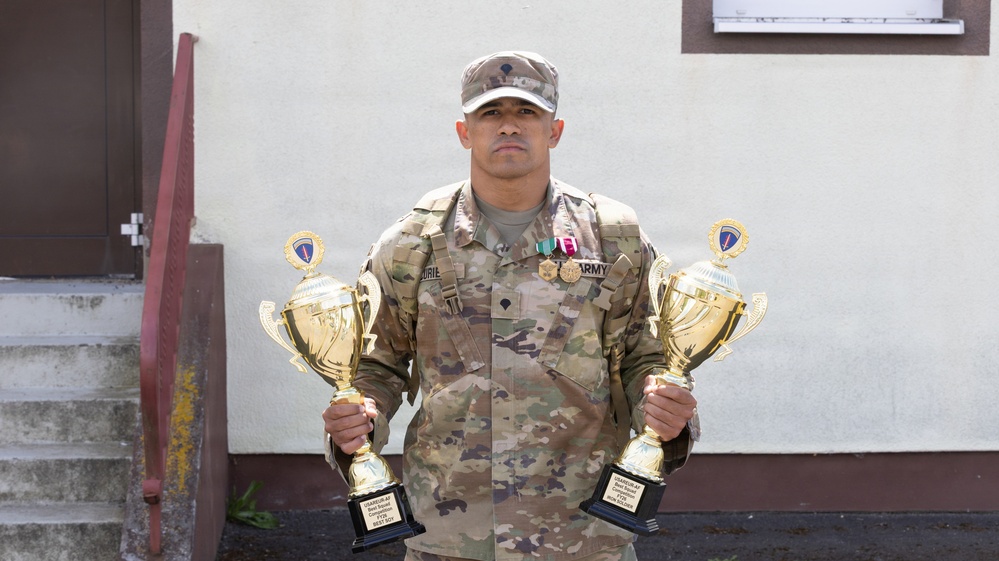 A U.S. Army soldier in multicam/OCP camouflage uniform stands holding two large gold trophies, one in each hand. He is wearing medals and ribbons on his chest, and his uniform displays 'ARMY' tape and a name tape. The soldier appears to be posing for an official or commemorative photograph in front of a building.