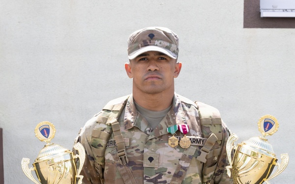 A U.S. Army soldier in multicam/OCP camouflage uniform stands holding two large gold trophies, one in each hand. He is wearing medals and ribbons on his chest, and his uniform displays 'ARMY' tape and a name tape. The soldier appears to be posing for an official or commemorative photograph in front of a building.