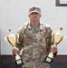 A U.S. Army soldier in multicam/OCP camouflage uniform stands holding two large gold trophies, one in each hand. He is wearing medals and ribbons on his chest, and his uniform displays 'ARMY' tape and a name tape. The soldier appears to be posing for an official or commemorative photograph in front of a building.