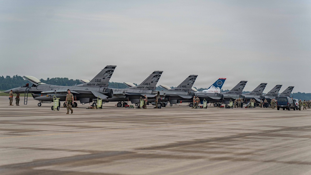 A row of F-16 Fighting Falcon jets belonging to the South Dakota Air National Guard are lined up on a flight line apron. Multiple ground crew personnel are visible performing maintenance and pre/post-flight servicing activities around the aircraft. One aircraft features a distinctive heritage tail marking with what appears to be a colonial-era flag design.