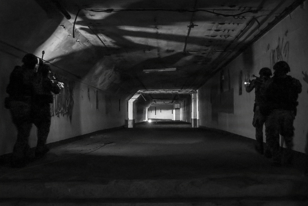 A black and white photograph showing two armed, helmeted figures in tactical gear positioned on opposite sides of a long corridor or tunnel. The corridor appears to be an abandoned or industrial space with graffiti on the walls and fluorescent lighting in the distance. The figures are in a tactical clearing posture, weapons raised toward the far end of the passage.