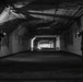 A black and white photograph showing two armed, helmeted figures in tactical gear positioned on opposite sides of a long corridor or tunnel. The corridor appears to be an abandoned or industrial space with graffiti on the walls and fluorescent lighting in the distance. The figures are in a tactical clearing posture, weapons raised toward the far end of the passage.