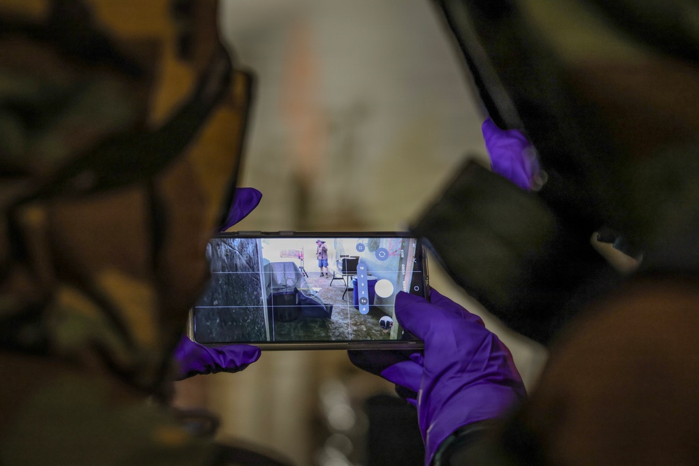 A person wearing purple latex/nitrile gloves and camouflage clothing is holding a smartphone horizontally, capturing a photo or video of an outdoor scene. The phone screen displays what appears to be a damaged or flooded area with a person standing in the background. The use of protective gloves suggests a potentially hazardous or forensic/emergency response context.