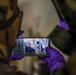 A person wearing purple latex/nitrile gloves and camouflage clothing is holding a smartphone horizontally, capturing a photo or video of an outdoor scene. The phone screen displays what appears to be a damaged or flooded area with a person standing in the background. The use of protective gloves suggests a potentially hazardous or forensic/emergency response context.