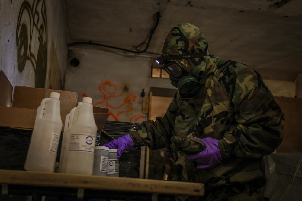 A person wearing full CBRN/hazmat protective gear, including a military camouflage suit, gas mask with headlamp, and purple nitrile gloves, is examining containers and boxes on a table inside a dilapidated building. The environment shows signs of abandonment with graffiti on the walls. The scene appears to depict a hazardous materials inspection or training exercise.