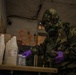 A person wearing full CBRN/hazmat protective gear, including a military camouflage suit, gas mask with headlamp, and purple nitrile gloves, is examining containers and boxes on a table inside a dilapidated building. The environment shows signs of abandonment with graffiti on the walls. The scene appears to depict a hazardous materials inspection or training exercise.