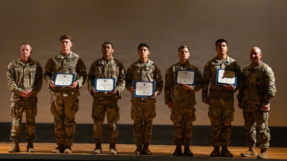 Seven military personnel in camouflage uniforms are standing in a line on a stage during what appears to be an awards or recognition ceremony. Five of the individuals are holding blue-bordered certificates or commendation documents. Two individuals on the far left and far right appear to be senior personnel presiding over the ceremony.