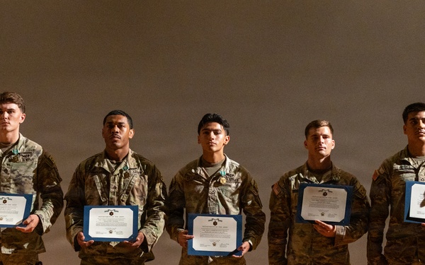 Seven military personnel in camouflage uniforms are standing in a line on a stage during what appears to be an awards or recognition ceremony. Five of the individuals are holding blue-bordered certificates or commendation documents. Two individuals on the far left and far right appear to be senior personnel presiding over the ceremony.