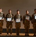 Seven military personnel in camouflage uniforms are standing in a line on a stage during what appears to be an awards or recognition ceremony. Five of the individuals are holding blue-bordered certificates or commendation documents. Two individuals on the far left and far right appear to be senior personnel presiding over the ceremony.