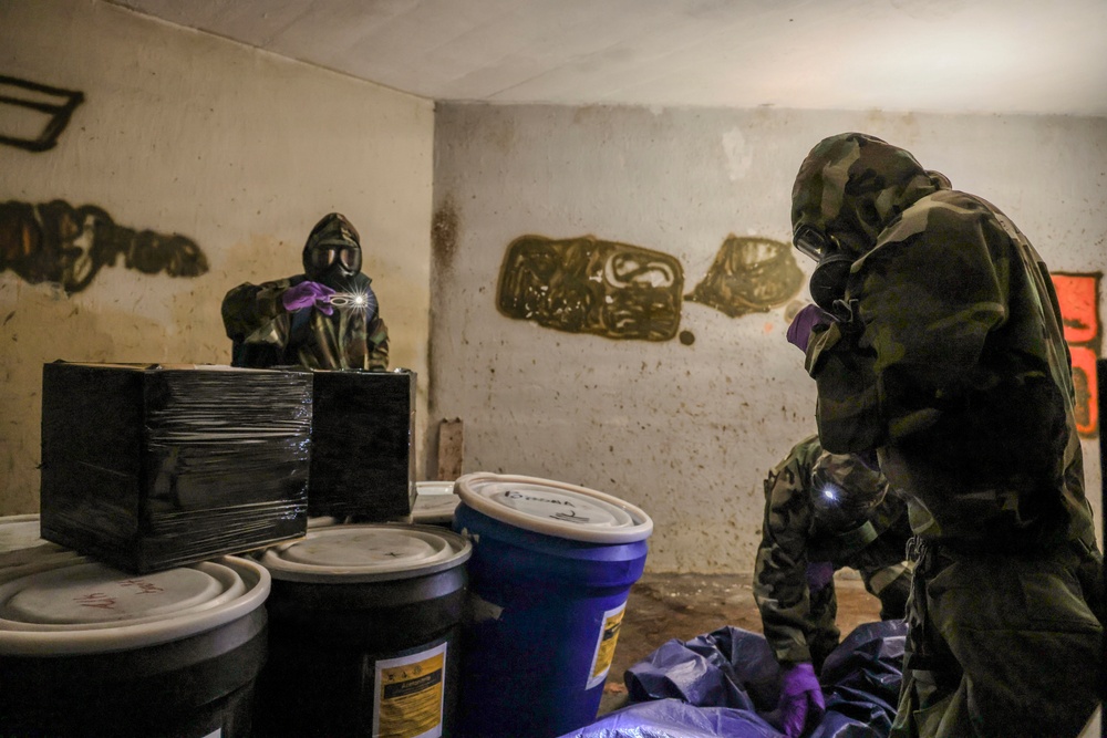The image shows personnel wearing full CBRN (Chemical, Biological, Radiological, Nuclear) protective gear including gas masks and camouflage hazmat suits operating inside a confined, graffiti-covered room. They are examining or cataloging containers including black-wrapped packages and plastic drums/barrels. The scene appears consistent with a law enforcement, military, or emergency response operation involving potentially hazardous materials.