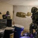 The image shows personnel wearing full CBRN (Chemical, Biological, Radiological, Nuclear) protective gear including gas masks and camouflage hazmat suits operating inside a confined, graffiti-covered room. They are examining or cataloging containers including black-wrapped packages and plastic drums/barrels. The scene appears consistent with a law enforcement, military, or emergency response operation involving potentially hazardous materials.