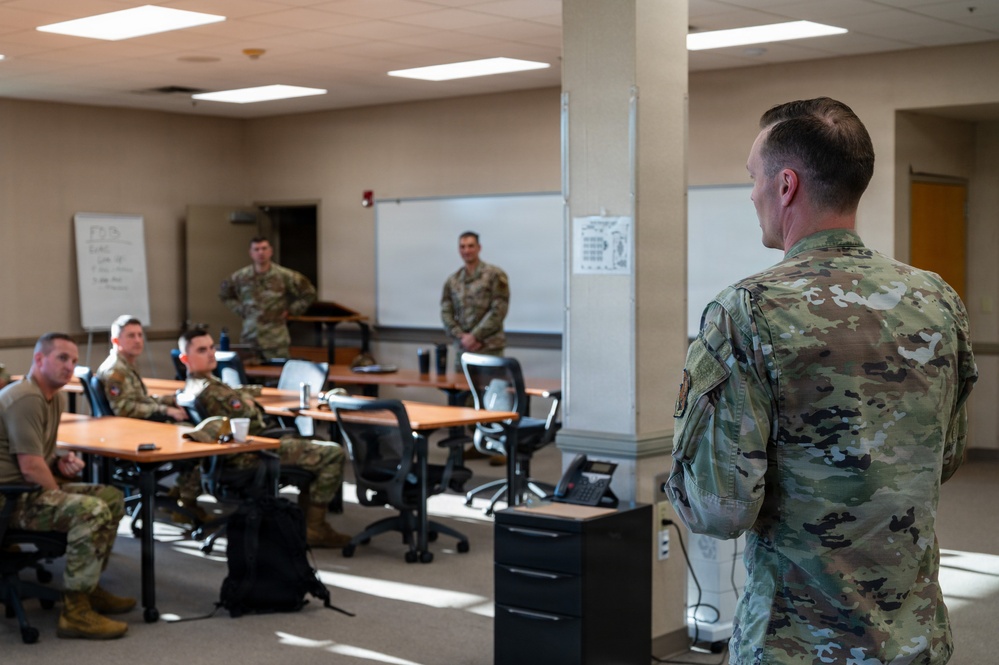 A military briefing or meeting is taking place in a classroom-style room where several uniformed personnel are gathered. One individual stands at the foreground addressing the group, while others are seated at tables or standing near a whiteboard at the back of the room. All personnel are wearing OCP (Operational Camouflage Pattern) uniforms.