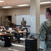 A military briefing or meeting is taking place in a classroom-style room where several uniformed personnel are gathered. One individual stands at the foreground addressing the group, while others are seated at tables or standing near a whiteboard at the back of the room. All personnel are wearing OCP (Operational Camouflage Pattern) uniforms.