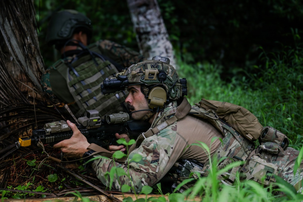 Two military personnel in full tactical gear are positioned in a prone/low stance at the base of a tree in a dense forest environment. The foreground soldier is equipped with advanced optics, night vision device, communications headset, and an assault rifle with multiple attachments. The image appears to depict a training exercise or tactical operation in a jungle or heavily vegetated area.