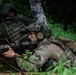 Two military personnel in full tactical gear are positioned in a prone/low stance at the base of a tree in a dense forest environment. The foreground soldier is equipped with advanced optics, night vision device, communications headset, and an assault rifle with multiple attachments. The image appears to depict a training exercise or tactical operation in a jungle or heavily vegetated area.
