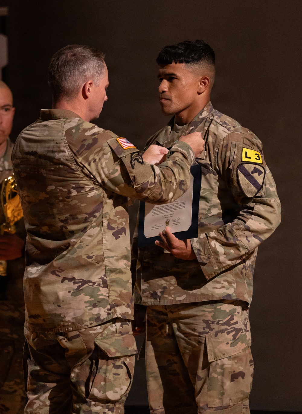 Two U.S. Army soldiers in Operational Camouflage Pattern (OCP) uniforms are engaged in what appears to be a military awards or pinning ceremony. An older soldier is pinning something onto the chest of a younger soldier, who is holding an official document or certificate in a blue folder. The younger soldier displays a 1st Cavalry Division patch and an 'L3' unit identifier on his left shoulder.