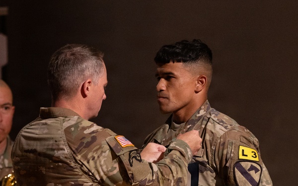 Two U.S. Army soldiers in Operational Camouflage Pattern (OCP) uniforms are engaged in what appears to be a military awards or pinning ceremony. An older soldier is pinning something onto the chest of a younger soldier, who is holding an official document or certificate in a blue folder. The younger soldier displays a 1st Cavalry Division patch and an 'L3' unit identifier on his left shoulder.