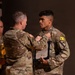 Two U.S. Army soldiers in Operational Camouflage Pattern (OCP) uniforms are engaged in what appears to be a military awards or pinning ceremony. An older soldier is pinning something onto the chest of a younger soldier, who is holding an official document or certificate in a blue folder. The younger soldier displays a 1st Cavalry Division patch and an 'L3' unit identifier on his left shoulder.
