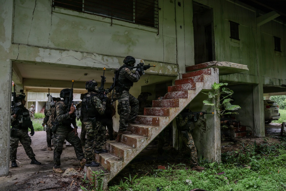 A group of armed military or law enforcement personnel in tactical gear are conducting what appears to be a building clearance or urban warfare training exercise at a dilapidated two-story structure. Multiple operators are stacked near a brick staircase, with the lead operator aiming a rifle toward the upper floor entrance. The building shows significant weathering and deterioration, consistent with a training facility or abandoned structure.
