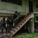 A group of armed military or law enforcement personnel in tactical gear are conducting what appears to be a building clearance or urban warfare training exercise at a dilapidated two-story structure. Multiple operators are stacked near a brick staircase, with the lead operator aiming a rifle toward the upper floor entrance. The building shows significant weathering and deterioration, consistent with a training facility or abandoned structure.