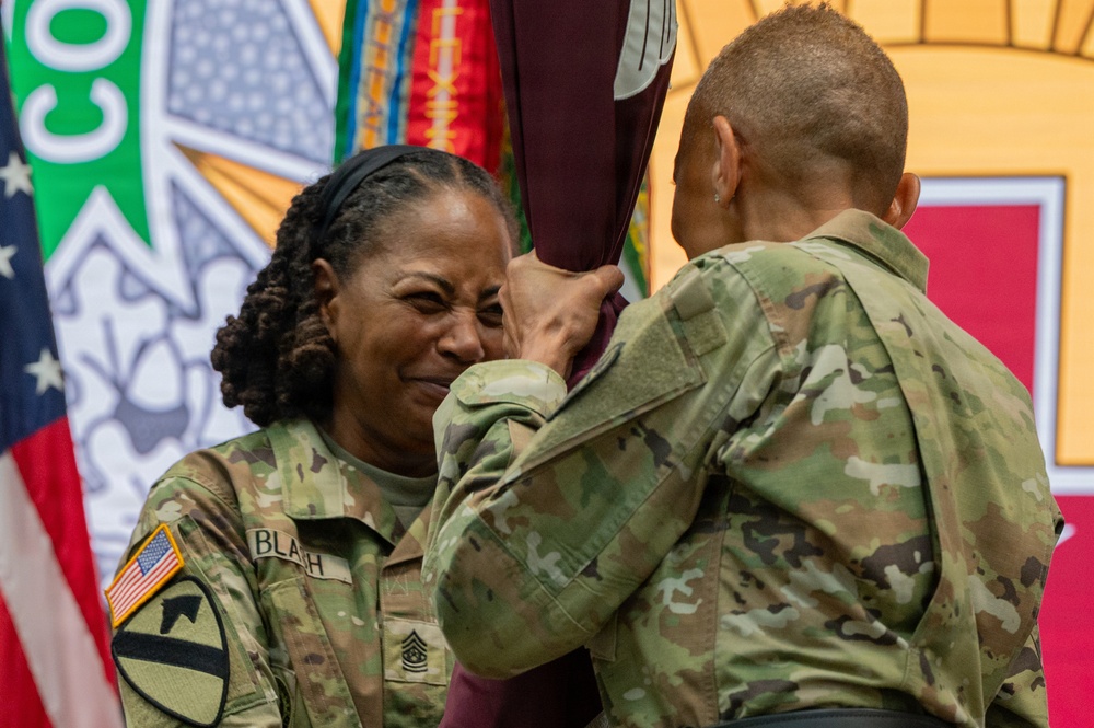 Two U.S. Army soldiers in MultiCam/OCP camouflage uniforms are participating in what appears to be a military ceremony, likely a change of command or change of responsibility. The soldier facing the camera, identified by the name tape 'BLAH' and wearing a Sergeant Major rank insignia, appears emotional while holding or receiving a unit guidon or flag. Military colors and flags are visible in the background.
