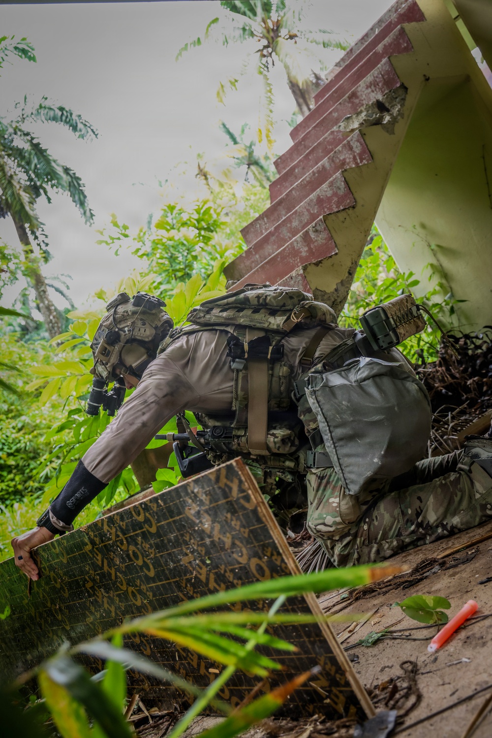 A heavily equipped individual in tactical/military gear is navigating through a dilapidated structure surrounded by dense tropical vegetation. The person is crouching and maneuvering around debris, concrete rubble, and a deteriorating staircase in what appears to be an abandoned building. The setting suggests a jungle or tropical environment, possibly during a training exercise or operational movement.