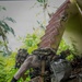 A heavily equipped individual in tactical/military gear is navigating through a dilapidated structure surrounded by dense tropical vegetation. The person is crouching and maneuvering around debris, concrete rubble, and a deteriorating staircase in what appears to be an abandoned building. The setting suggests a jungle or tropical environment, possibly during a training exercise or operational movement.