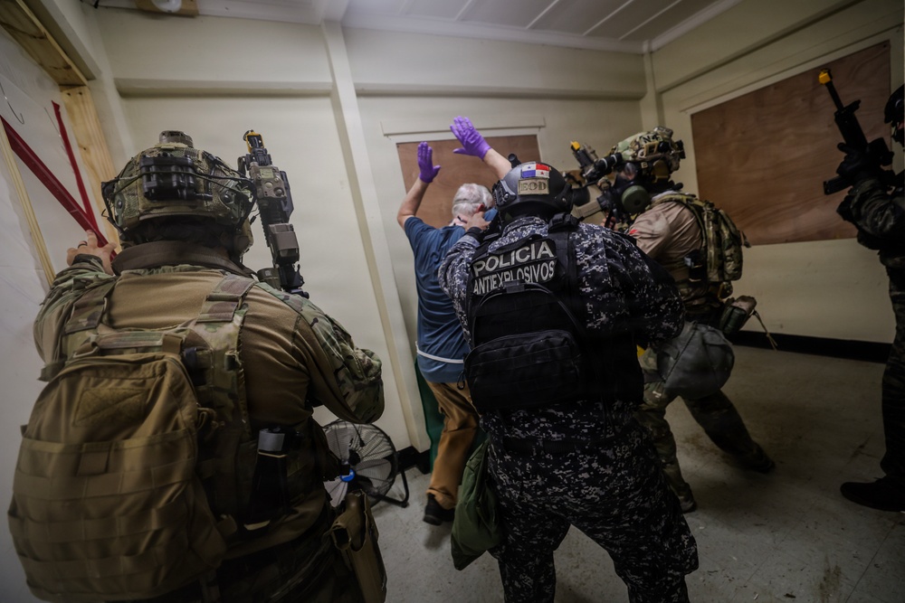 The image depicts what appears to be a law enforcement or military training exercise involving tactical personnel. Officers wearing 'Policia Antiexplosivos' (Anti-Explosives Police) vests, along with heavily equipped tactical operators, are detaining or controlling a civilian-dressed individual against a wall. The Panamanian flag patch is visible on at least one officer's uniform, suggesting this involves Panamanian security forces.