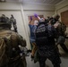 The image depicts what appears to be a law enforcement or military training exercise involving tactical personnel. Officers wearing 'Policia Antiexplosivos' (Anti-Explosives Police) vests, along with heavily equipped tactical operators, are detaining or controlling a civilian-dressed individual against a wall. The Panamanian flag patch is visible on at least one officer's uniform, suggesting this involves Panamanian security forces.