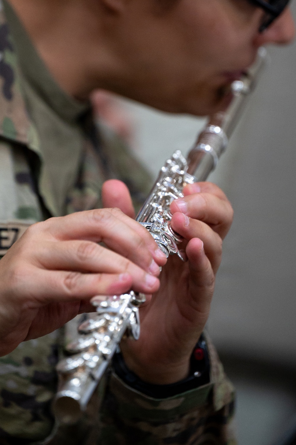 A person wearing a military camouflage uniform is playing a silver flute. The image is focused on the hands and the instrument, with the player's face blurred in the background. The silver keys and body of the flute are sharply rendered in the foreground.