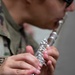 A person wearing a military camouflage uniform is playing a silver flute. The image is focused on the hands and the instrument, with the player's face blurred in the background. The silver keys and body of the flute are sharply rendered in the foreground.
