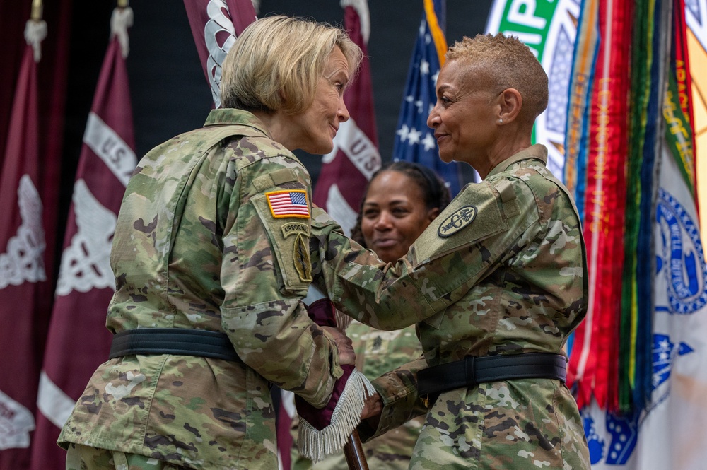 Two female U.S. Army soldiers are engaged in what appears to be a formal military ceremony, likely a change of command or award presentation, as one soldier reaches toward the other's chest area. Both wear Army Combat Uniforms (ACU) in MultiCam/OCP pattern with black belts. Multiple military unit flags and colors are displayed in the background.