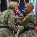 Two female U.S. Army soldiers are engaged in what appears to be a formal military ceremony, likely a change of command or award presentation, as one soldier reaches toward the other's chest area. Both wear Army Combat Uniforms (ACU) in MultiCam/OCP pattern with black belts. Multiple military unit flags and colors are displayed in the background.