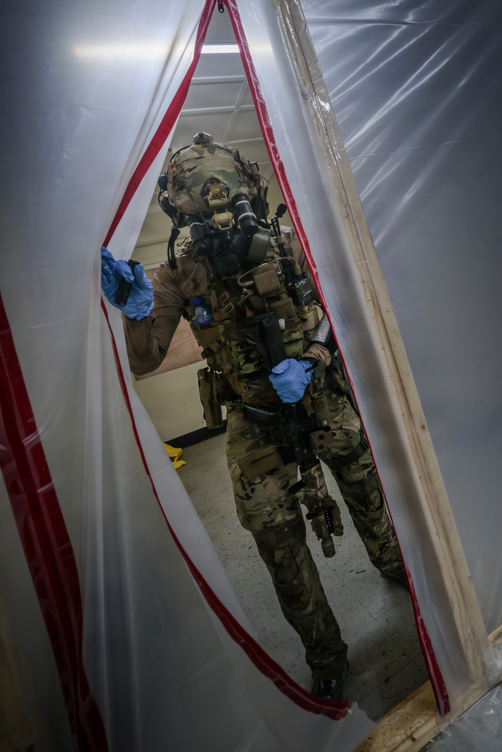A heavily equipped military or law enforcement operator in full tactical gear is passing through a plastic sheeting barrier with red zipper trim, consistent with a CBRN (Chemical, Biological, Radiological, Nuclear) decontamination corridor or containment zone. The individual is wearing a full-face gas mask, multicam camouflage uniform, plate carrier with accessories, and blue nitrile gloves. A second similarly equipped individual is partially visible in the background.