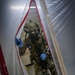 A heavily equipped military or law enforcement operator in full tactical gear is passing through a plastic sheeting barrier with red zipper trim, consistent with a CBRN (Chemical, Biological, Radiological, Nuclear) decontamination corridor or containment zone. The individual is wearing a full-face gas mask, multicam camouflage uniform, plate carrier with accessories, and blue nitrile gloves. A second similarly equipped individual is partially visible in the background.