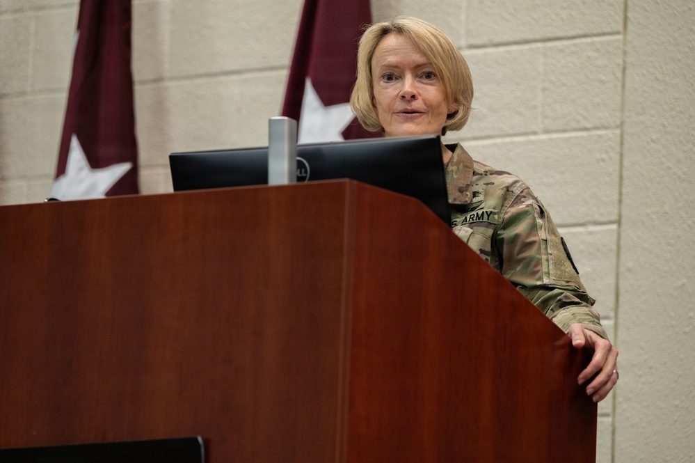 A U.S. Army soldier in Operational Camouflage Pattern (OCP) uniform stands at a wooden podium with a Dell monitor, appearing to deliver a briefing or speech. Behind the individual, a flag or banner with a white star on a dark red/maroon background is visible, suggesting a military headquarters or command setting. The 'U.S. ARMY' tape is clearly visible on the uniform.