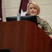 A U.S. Army soldier in Operational Camouflage Pattern (OCP) uniform stands at a wooden podium with a Dell monitor, appearing to deliver a briefing or speech. Behind the individual, a flag or banner with a white star on a dark red/maroon background is visible, suggesting a military headquarters or command setting. The 'U.S. ARMY' tape is clearly visible on the uniform.