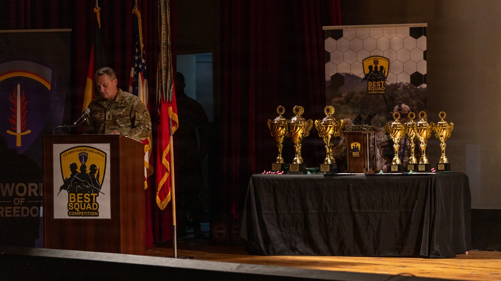 A military officer in camouflage uniform speaks at a podium during the U.S. Army Europe and Africa 'Best Squad Competition' ceremony. Multiple gold trophies are displayed on a draped table to the right of the podium, alongside a branded banner for the competition. The event appears to be an awards ceremony held on a stage with military flags and decorative backdrops.