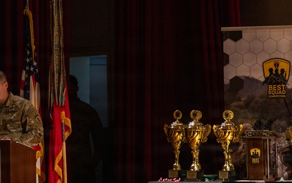 A military officer in camouflage uniform speaks at a podium during the U.S. Army Europe and Africa 'Best Squad Competition' ceremony. Multiple gold trophies are displayed on a draped table to the right of the podium, alongside a branded banner for the competition. The event appears to be an awards ceremony held on a stage with military flags and decorative backdrops.