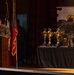 A military officer in camouflage uniform speaks at a podium during the U.S. Army Europe and Africa 'Best Squad Competition' ceremony. Multiple gold trophies are displayed on a draped table to the right of the podium, alongside a branded banner for the competition. The event appears to be an awards ceremony held on a stage with military flags and decorative backdrops.