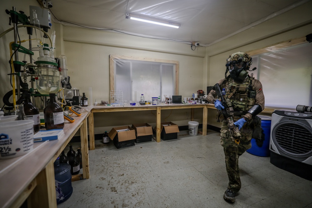 A fully equipped tactical operator wearing military multicam uniform, gas mask, and blue nitrile gloves is standing inside what appears to be a clandestine chemical laboratory. The room contains extensive chemical apparatus, reagent bottles, glassware, and laboratory benches. This appears to be a law enforcement or military raid/exploitation of an illicit drug manufacturing facility.