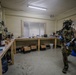 A fully equipped tactical operator wearing military multicam uniform, gas mask, and blue nitrile gloves is standing inside what appears to be a clandestine chemical laboratory. The room contains extensive chemical apparatus, reagent bottles, glassware, and laboratory benches. This appears to be a law enforcement or military raid/exploitation of an illicit drug manufacturing facility.
